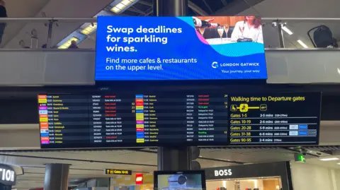 Two departure boards at Gatwick airport on 28 December, showing status of flights, some of which are delayed. A third board shows the walking time to reach various departure gates. In the background, there are two stores - a Lego store and a Hugo Boss store.