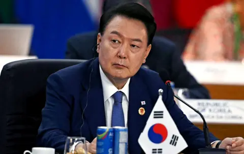 AFP President Yoon Suk Yeol looks away from camera while wearing a suit and tie with an earpiece in, with a miniature South Korea flag and refreshments on the table in front of him as he sits during the third session of the G20 Leaders' Meeting in Rio de Janeiro, Brazil, on November 19, 2024.