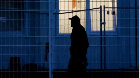 EPA A man in a baseball cap walks past a window at night. He's silhouetted against the building and the soft white light coming from the window.
