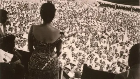 Herbert Weiss Wearing a patterened dress, Andrée gives a speech to a large crowd of men and women at a rally in the Democratic Republic of Congo