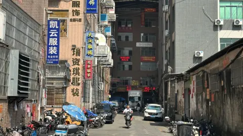 Xiqing Wang/BBC A man on a motorbike drives down a street with buildings on either side 