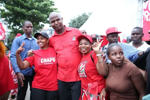 AFP Daniel Chapo celebrates his victory with supporters in Maputo in December. They are wearing red, the colour of his Frelimo party. Two woman put their arms around the politician and make the 'peace' sign with their hands.