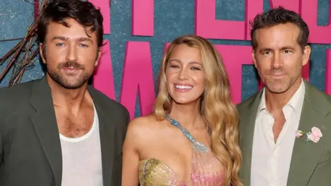 Getty Images Three people - Brandon Sklenar, Blake Lively and Ryan Reynolds - are dressed in formal attire and posing for a photo. They are stood in front of a large sign that reads "It Ends With Us"