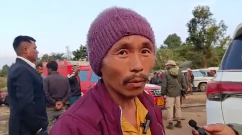 Dilip Sharma A photo of Ravi Rai, wearing a red hat, a red jacket and a yellow T-shirt, speaking to a reporter. He is among the miners that successfully escaped when an illegal mine was flooded in India's Assam state.