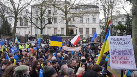 PA Media A group of protesters - holding Ukraine flags and a poster reading "Alexei Navalny 04/06/1976-16/02/2024 The bravest of the brave, We will not forget - outside the Russian embassy in London - a grand white building with a Russian flag flying over it's door.