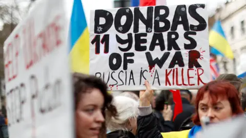 Reuters Two women stand in a crowd of protesters, one holding up a poster saying "Donbas 11 years of war, Russia is a killer"