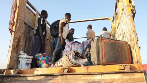 UNHCR A group of men are standing in the back of an open lorry, a suitcase, a rolled-up carpet and some other luggage can be seen in the foreground at the edge of the back of the vehicle.