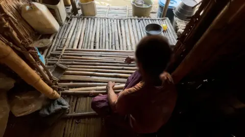 BBC Image showing Zeya with his back to the camera, sitting cross legged on a floor made of bamboo poles, holding a cigarette in his right hand. His head is blurred and there are several water containers in buckets in the background. 