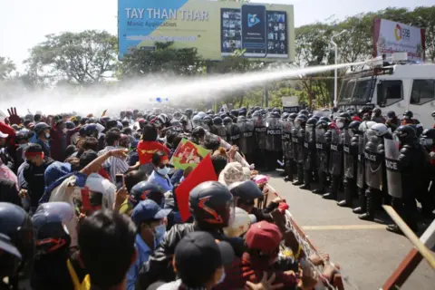 Getty Images A line of police with riot shields and helmets face a crowd of protesters with a gap of a few metres between the two sides. Behind the police is a white vehicle spraying water from a water cannon on its roof, as protesters demonstrate against the 1 Feb military coup in the Myanmar capital Naypyidaw on 9 Feb, 2021