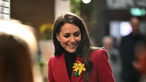 Ben Stansall/PA Wire The Princess of Wales is smiling as she looks down, she is wearing a red coat and is holding a pink and white polka dot tube. Catherine has a large yellow daffodil broach attached to her red coat. She is in focus and the foreground is people waiting to meet her, their heads are out of focus.
