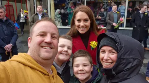 Gavin Hailes Selfie of Gavin Hailes, 41 who is wearing a mustard yellow hoodie, his son Ralphie, 12, who is wearing a black jumper and has short spiked hair, Joseph, 8, who is wearing a green Puffa jacket, and aunt Hannah Jones, 45. Princess Catherine smiles behind them. 