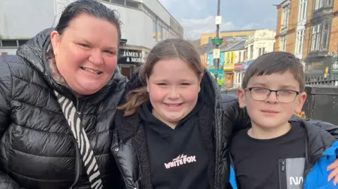 Lisa Maunder, 46, stands on the left smiling at the camera, she has a black Puffa jacket and a zebra print bag strap across her shoulder. She has her hair pulled back in a ponytail. Daughter Darcy, 10, stands next to her, she is wearing a White Fox jumper which is black and has pink text, under a black Puffa jacket with black Sherpa lining, she has her long hair pulled into a ponytail. Quade, 8, stands next to her wearing a black t-shirt and a blue raincoat. He has short hair and is wearing glasses.