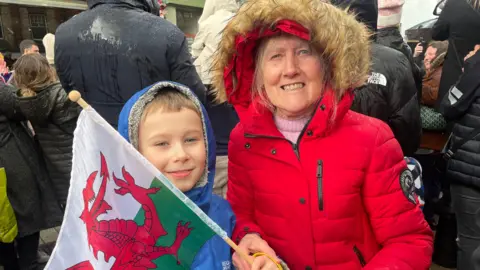 Patricia Thomas stands on the right wearing a red Puffa jacket and a hood with fur on. She is holding a Welsh flag. To her left is her grandson Harrison who is wearing a blue raincoat, he has short blonde hair visible beneath his hood.