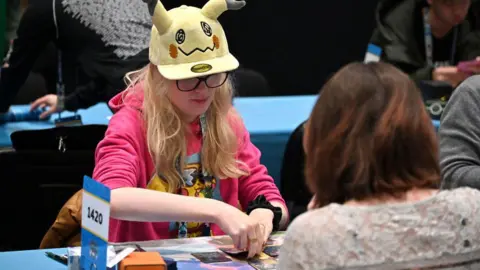 Getty Images A young Pokémon fan sits across a table from an opponent during a match of the trading card game. Wearing a yellow baseball cap with Pikachu ears and a bright pink hoody with a colourful Pikachu print on it, she looks thoughtful as she considers her next move. 