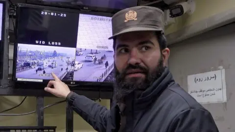 A man in a hat points to a surveillance monitor while looking towards the camera