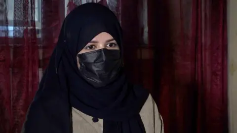 A woman wearing a headscarf and face mask sits in front of the camera