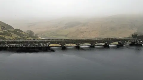 BBC An aerial photo of the imposing Claerwen Reservoir. Its dam is made from grey stone and has arches along the top. It is surrounded by barren misty hills.