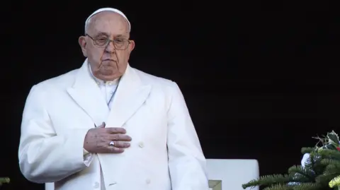 Reuters Wearing a white coat the Pope touches his chest while standing up and looking down at a crowd on Christmas Day