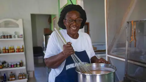 Gemma Handy Novella Payne, wearing glasses and a hair covering, stirs a pot containing a sauce she makes using traditional recipes