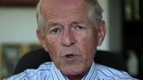 BBC John Smyth QC looks on during a court hearing. He's sat on a chair and wearing a stripy shirt.