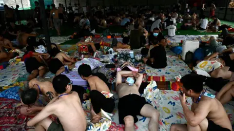 Getty Images A roomful of men, some lying down on mattreses, others sitting up or standing, in a makeshift camp. Many of them are shirtless and in shorts. 