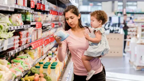 Getty Images A woman checks the price of an item in a grocery store while holding a baby