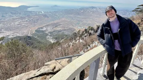 Joe Smith A man in black trousers, black jeans and purple fleece stands in front of a stone fence by a viewing platform. Behind him you can see mountains, trees and the sea