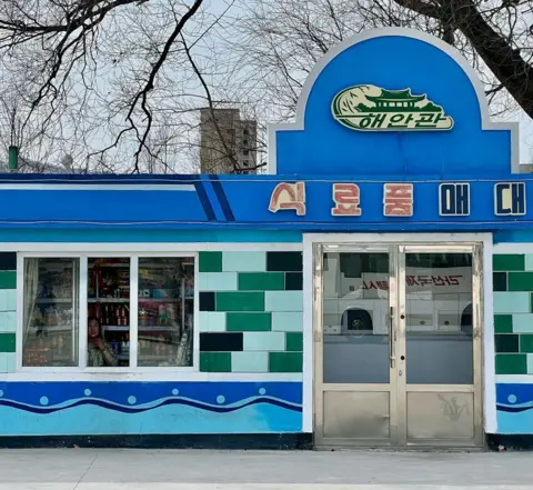 Joe Smith A photo of the exterior of a blue food store in North Korea. The exterior has green titles and there is a woman looking out of a window