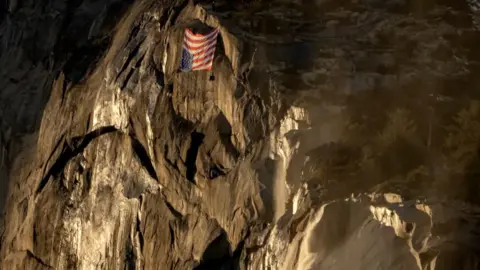 Photo by: Brittany Colt, www.brittanycolt.com, @brittanycolt Un upside down American Flag hangs on the face of a rock formation at dusk