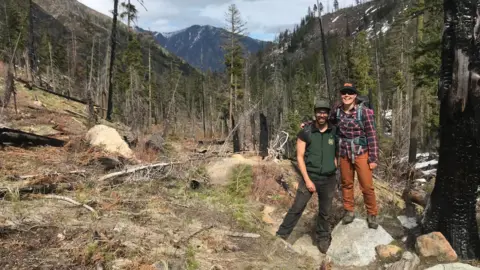 Submitted to BBC Demetrios and Thompson stand smiling in the middle of a clearing in a hilly forest, with a mountain peak visible in the background. Demetrios has a beard and is wearing a green sport vest and brown work pants and brown hiking boots, with a baseball cap shielding his eyes. Thompson is standing next to him on a rock so that she is taller, wearing orange work pants, a red flannel shirt, a baseball hat and a hiking backpack. 