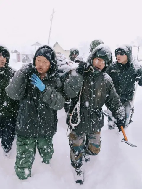 Indian Army via Reuters Two rescuers from the Indian Army, dressed in green overalls, carry items over their shoulders as they trek through heavy snow
