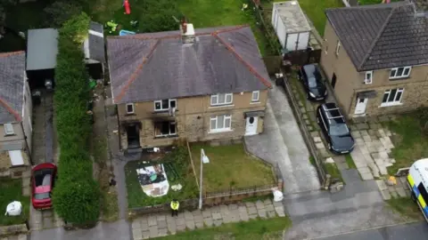 Spencer Stokes/BBC A burnt-out house, pictured from above. The front door and windows are covered with black fire damage and a police officer stands outside the property.