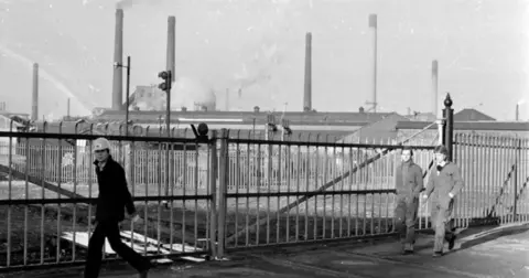 Getty Images A black and white photo shows three men walking past metal gates. Behind them are various smoking chimneys of the Corby steelworks. 