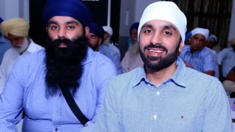 Gupreet Singh Johal Gurpreet and Jagtar Singh Johal sit in a room with other Sikh men. Gurprret is wearing a royal blue turban and a light blue shirt and has a long, dark beard. Jagtar is smiling and wearing a white turban and a blue and white checked shirt.