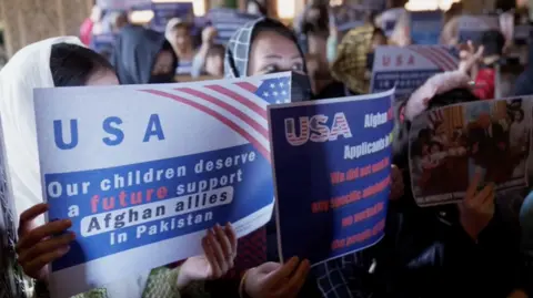 A small crowd of Afghan women hold up signs saying "USA, Our children deserve a future. Support Afghan allies in Pakistan." Another carries a photo of Donald Trump in the Oval Office. 