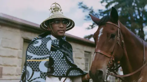 Getty Images Moshoeshoe II wears glasses, a traditional blanket and mokorotlo . He holds a horse by its reins.