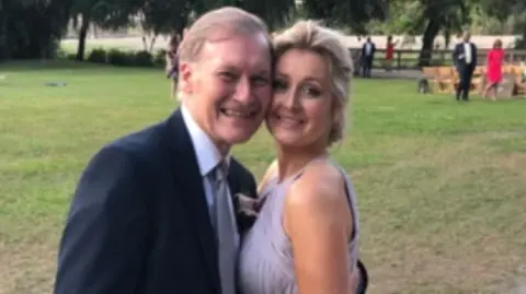 Katie Amess Sir David and Katie Amess standing on a grass lawn. Sir David is wearing a suit and Katie is wearing a sleeveless lilac dress. They are both smiling.