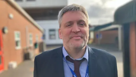 A man with a blue shirt and red tie stands in front of a school yard looking straight at the camera. 