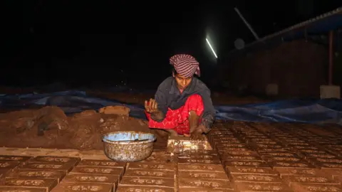 Jayaraj S A woman wearing a red dress and grey shirt is making brown coloured bricks. There is a pile of brown mix on her side which she uses to make bricks.