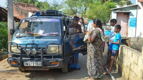 Gopika Lakshmi M A man wearing a brown shirt and blue cloth tied around his waist hands cash to a female customer who is wearing a grey night dress. The man stands alongside his blue van. 