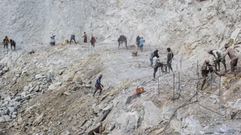 Mukesh K A landscape shot of a vast quarry showing numerous workers hard at work drilling, with yellow power cables snaking across the ground