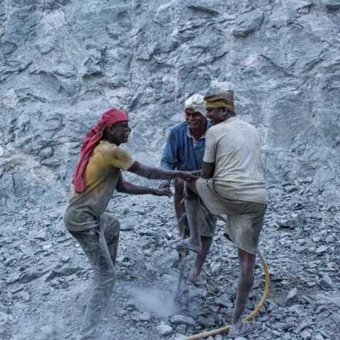 Mukesh K A trio of quarry workers, wearing yellow, blue and white T-shirts, grin at each other and chat as they drill the ground