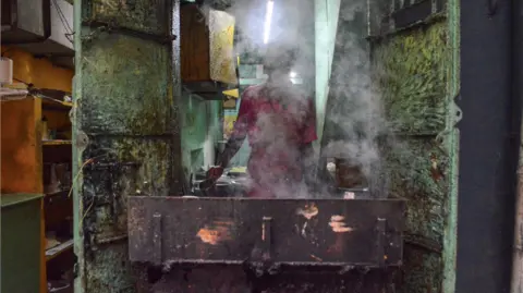 Govarthanan L S A man in a pink shirt cooks food on a hot open stove, flanked by grimy steel doors, shrouded in steam