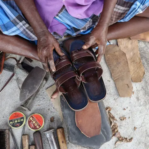 Saran R A close-up image of a cobbler's hands holding a pair of maroon leather sandals, surrounded by the tools of his trade