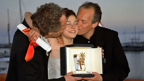 Getty Images Émilie Dequenne holding a Cannes award with Luc and Jean-Pierre Dardenne on either side kissing her on both cheeks in 1999