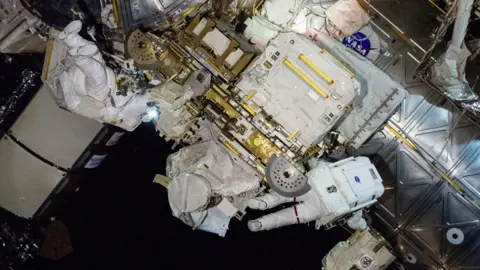 NASA Butch and Suni float amidst highly technical equipment outside of the ISS, wearing white space suits. You can't tell who is who as one has their back to the camera, the other has their feet in the air. 