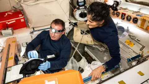 NASA Butch and Suni carry out an experiment - Butch wears a headlight and blue surgical gloves. He is sitting in a type of pit and appears to be looking inside a black bag. Suni hangs just over him, looking at what he is doing. 