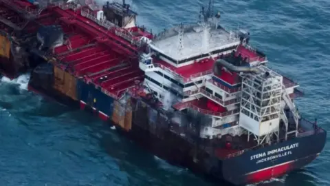 Reuters A large oil tanker with a gaping hole in its port (left) side. The area around the bridge, at the stern of the ship, is blackened from fire. 