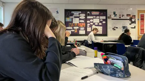 BBC There are a row of students in a classroom in navy jumpers with their heads down looking at their notebooks. A pencil case is on the table. 