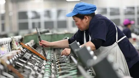 Getty Images A female worker works on a spun silk production line at a Chinese factory. She is wearing a blue cap and a white apron over her navy blue uniform. 